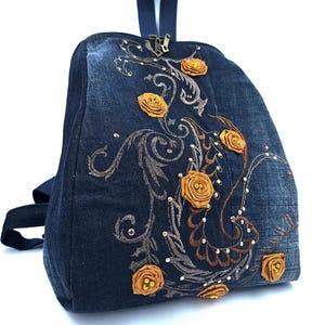 May include: A dark blue denim backpack featuring an embroidered swirling pattern in brown and metallic thread. The bag is decorated with several three-dimensional mustard yellow fabric rosettes with small beads at their centers and metallic studs.