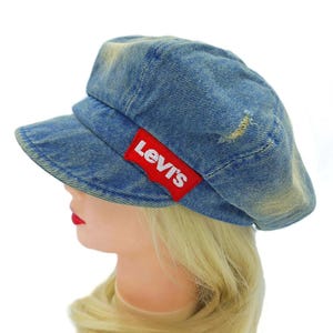 May include: A blue denim cap with a short brim and a red Levi's logo patch. The cap has a distressed look with faded areas and a small tear. The cap is worn by a mannequin with blonde hair and red lipstick.