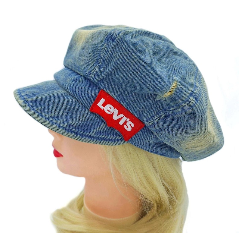 May include: A blue denim cap with a short brim and a red Levi's logo patch. The cap has a distressed look with faded areas and a small tear. The cap is worn by a mannequin with blonde hair and red lipstick.