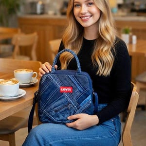 May include: A square-shaped denim handbag featuring a patchwork design of various blue denim fabric strips. The bag includes a top handle, a shoulder strap, and a prominent red rectangular Levi's brand label centered on the front exterior.