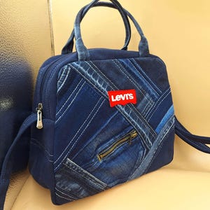 May include: A dark blue denim handbag featuring a patchwork design with diagonal stitching. The bag has two top handles, a zippered front pocket, and a red Levi's brand patch centered on the front. The exterior is constructed from various shades of blue denim fabric with visible seams.