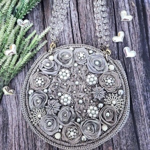 Può includere: Una borsa rotonda grigia con una tracolla a catena trasparente. La borsa è decorata con rose di tessuto grigio, perle finte e abbellimenti di perline. La borsa è ambientata su uno sfondo di legno con cuori decorativi e verde.