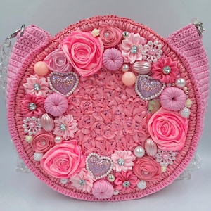 Könnte beinhalten: Eine rosa, runde Handtasche mit gewebter Textur, verziert mit Stoffblumen, Perlen und herzförmigen Verzierungen. Die Tasche hat einen Kettenriemen und ohrenförmige Akzente, die ein skurriles Design schaffen.