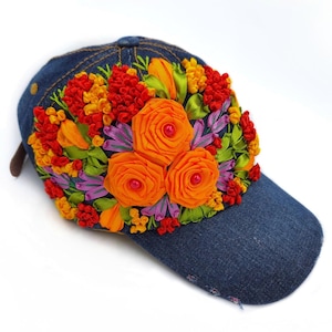 Könnte beinhalten: Eine dunkelblaue Baseballkappe aus Jeansstoff mit einer aufwendigen dreidimensionalen Blumenstickerei auf der Vorderseite. Das Design umfasst leuchtend orangefarbene Bandrosen, rote und gelbe Texturcluster.