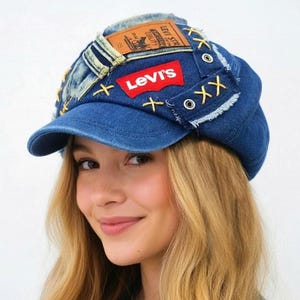 Puede incluir: Una gorra de estilo newsboy en mezclilla azul con un parche rojo del logotipo de Levi's, una etiqueta color canela estilo cuero, bordes deshilachados y detalles decorativos de punto de cruz amarillo. La gorra tiene paneles de mezclilla y ojales.