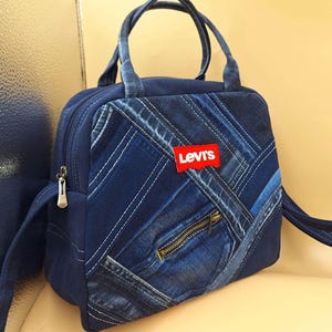 May include: A blue denim handbag featuring a patchwork design of various denim shades with diagonal stitching. The bag includes two top handles, a front zippered pocket, and a red rectangular Levi's brand patch centered on the front panel.