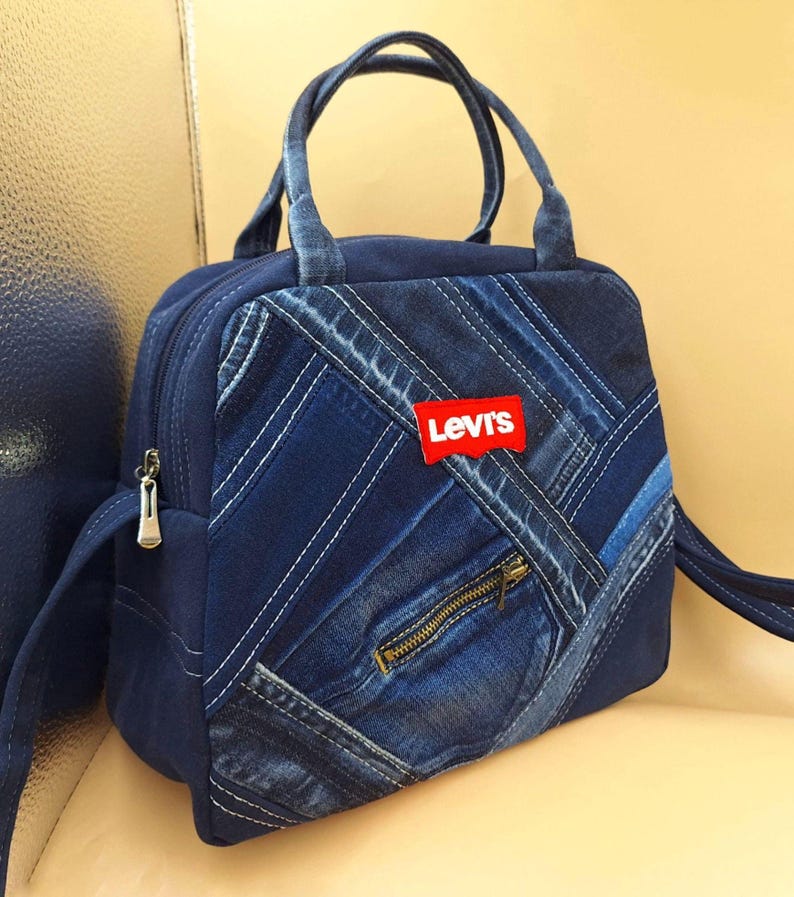 May include: A blue denim handbag featuring a patchwork design of various denim shades with diagonal stitching. The bag includes two top handles, a front zippered pocket, and a red rectangular Levi's brand patch centered on the front panel.