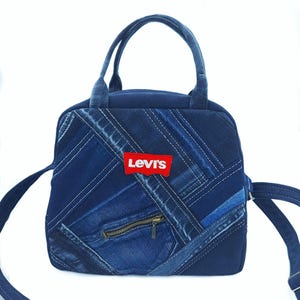 May include: A blue denim handbag featuring a patchwork design of various denim shades arranged in diagonal strips. The bag includes two top handles, a shoulder strap, a brass-colored zipper pocket, and a red Levi's brand label on the front.