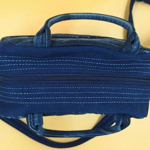 May include: A dark blue denim handbag featuring a rectangular shape with a top zipper closure. The bag includes two short handles and a long shoulder strap, with horizontal white stitching details across the fabric surface and sides.