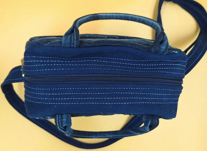May include: A dark blue denim handbag featuring a rectangular shape with a top zipper closure. The bag includes two short handles and a long shoulder strap, with horizontal white stitching details across the fabric surface and sides.