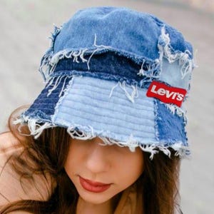 May include: A blue denim bucket hat constructed from various shades of patchwork denim fabric with frayed, raw edges throughout. The hat features a prominent red rectangular patch with the white text Levi's stitched onto the side panel.