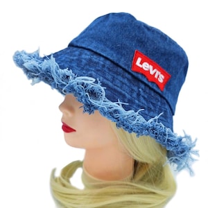 May include: A blue denim bucket hat with a frayed brim. The hat features a red Levi's logo patch. The hat is displayed on a mannequin head with blonde hair and red lipstick.