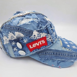 Puede incluir: Gorra de béisbol de mezclilla azul con diseño de patchwork. La gorra presenta un parche rojo con el logo de Levi's, detalles de encaje blanco y botones blancos. Hecha de tela vaquera.