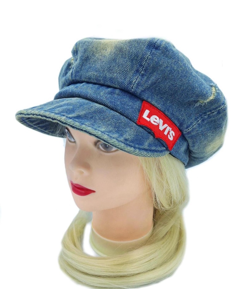 May include: A blue denim cap with a short brim. The cap has a distressed look with a red Levi's logo patch on the side. The cap is displayed on a mannequin head with blonde hair and red lipstick.