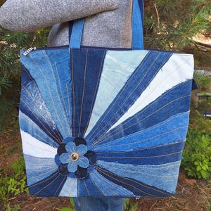 May include: A tote bag constructed from various shades of blue denim fabric arranged in a sunburst pattern. The bag features a central denim flower appliqué with a dark bead at its center, a top zipper closure, and two blue denim shoulder straps.