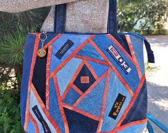 May include: A tote bag constructed from patchwork denim in various shades of blue, featuring orange fabric strips between the panels. The bag includes several rectangular brand labels, a small square leather tag labelled Hand Made, and a circular evil eye charm.