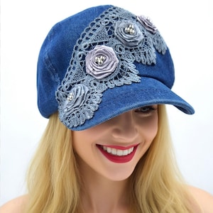 May include: A blue denim baseball cap featuring a decorative panel of grey lace trim arranged in a diagonal pattern. The lace is adorned with three grey fabric rosettes, each centered with a cluster of small, shiny silver-toned metallic beads.