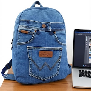 Peut inclure: Un sac à dos en jean bleu doté d'une poche avant avec un écusson en cuir marron de la marque Wrangler et des surpiqûres en W. Le design intègre des éléments de jean, notamment une ceinture, des rivets métalliques et des poches latérales.