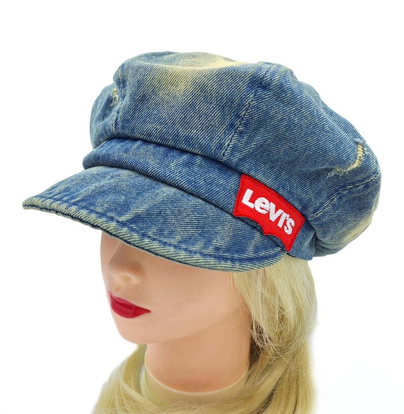May include: A blue denim newsboy cap with a short brim. The cap features a red Levi's logo patch on the side. The denim has a faded, worn look.