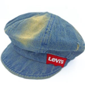 May include: A blue denim cap with a short brim and a rounded crown. The cap has a faded, worn appearance with a red Levi's logo patch on the side. The hat is a classic style.