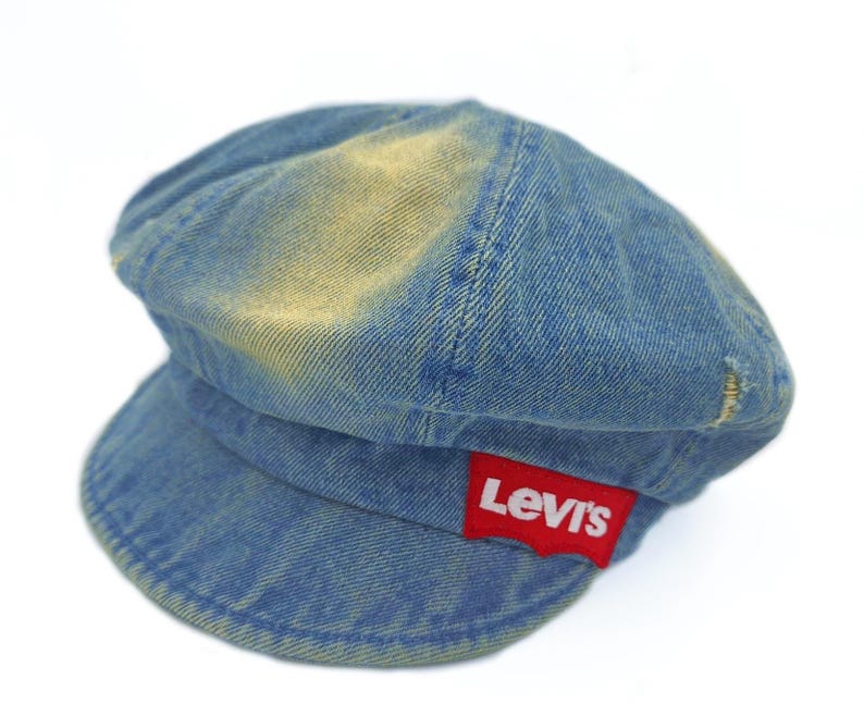 May include: A blue denim cap with a short brim and a rounded crown. The cap has a faded, worn appearance with a red Levi's logo patch on the side. The hat is a classic style.