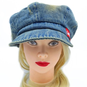 May include: A blue denim newsboy cap with a short brim. The cap has a distressed look with faded areas and a small red tag on the side. The cap is worn by a mannequin with blonde hair and red lipstick.