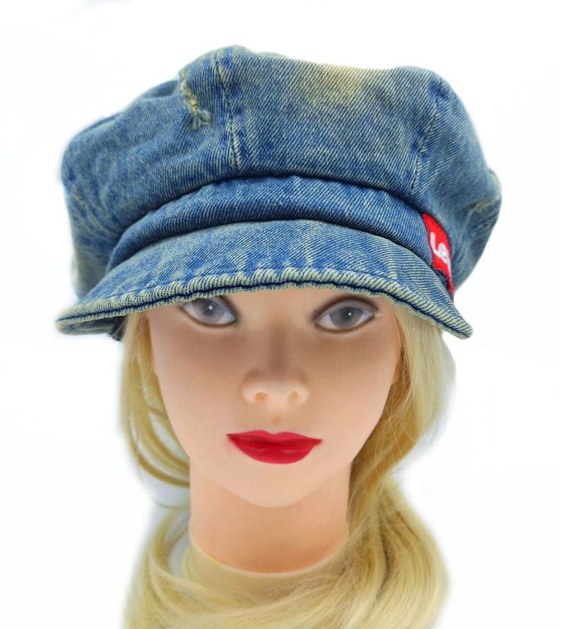 May include: A blue denim newsboy cap with a short brim. The cap has a distressed look with faded areas and a small red tag on the side. The cap is worn by a mannequin with blonde hair and red lipstick.