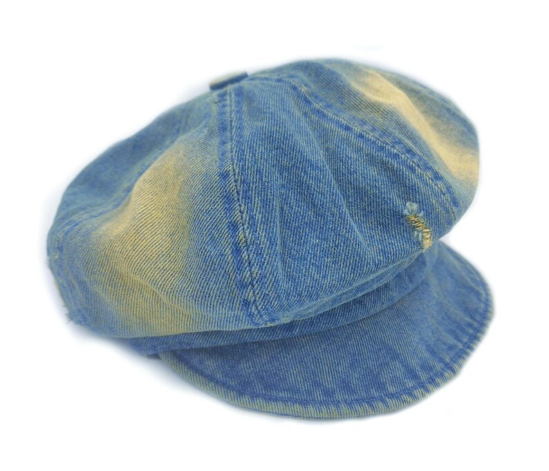 May include: A blue denim newsboy cap with a slightly faded, distressed appearance. The cap features a rounded crown, a short brim, and visible stitching details. The denim has a worn look with lighter patches.