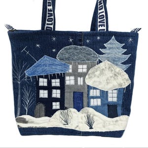 Könnte beinhalten: Jeans-Tote-Tasche mit einer Winterdorf-Szene. Die Tasche hat applizierte Häuser, einen Baum und Schnee mit weißen Akzenten. Die Griffe sind dunkelblau mit dem Text "AND LOVE U" in Weiß. Die Tasche ist aus dunkelblauem Jeansstoff.
