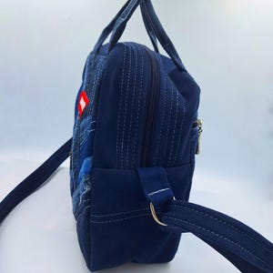 May include: A dark blue denim backpack featuring a patchwork design on the front panel, two top handles, and adjustable shoulder straps. The side panel shows a vertical zipper closure, white contrast stitching, and a small red rectangular tag.