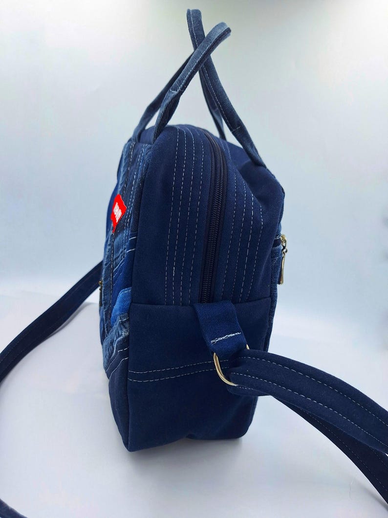 May include: A dark blue denim backpack featuring a patchwork design on the front panel, two top handles, and adjustable shoulder straps. The side panel shows a vertical zipper closure, white contrast stitching, and a small red rectangular tag.