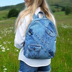 Puede incluir: Una mochila de mezclilla azul confeccionada con tela de jeans reciclada. La bolsa cuenta con múltiples bolsillos exteriores con cierres de botón, detalles de costuras diagonales y un asa superior. El diseño incorpora bolsillos originales.