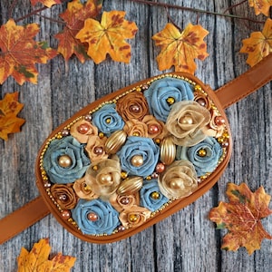 Può includere: Un marsupio marrone con un pannello frontale decorativo ornato da rose in tessuto nei toni del blu e del marrone chiaro, accentuato da perline dorate e ciondoli metallici ovali. La borsa ha una tracolla marrone liscia e uno scomparto ovale.