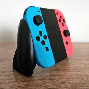 Peut inclure: Une console Nintendo Switch avec des manettes Joy-Con bleues et rouges. Les manettes sont fixées au corps noir de la console. La console repose sur un support noir. La Switch est sur une surface en bois.