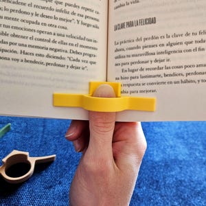May include: A yellow plastic thumb page holder is used to hold a book open. The book is open to a page with text in Spanish about forgiveness and happiness. The page holder is designed to fit over the thumb and has a curved shape to hold the page in place.