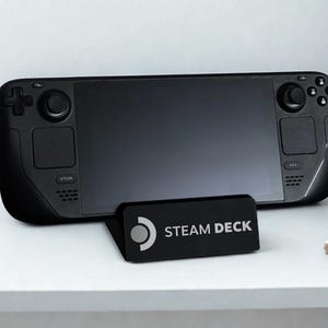 Pode incluir: Uma consola de jogos portátil preta com um ecrã grande, joysticks duplos e botões direcionais. O dispositivo é suportado por um suporte preto inclinado com o logótipo branco da Steam e o texto STEAM DECK impresso na parte frontal.