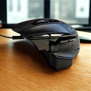 May include: A black computer mouse with a grey cable rests on a wooden surface. The mouse features a curved design with a scroll wheel and several buttons. The Logitech logo is visible. This mouse is designed for gaming.