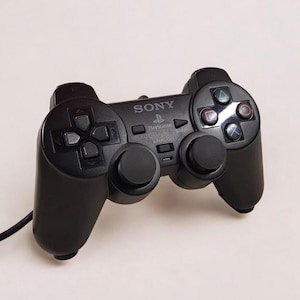 Może przedstawiać: Czarny kontroler Sony PlayStation 2 z połączeniem przewodowym. Kontroler posiada pad kierunkowy, drążki analogowe i przyciski akcji. Logo Sony jest widoczne na kontrolerze.