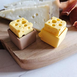 Puede incluir: Dos pequeños objetos amarillos con forma de queso y agujeros se asientan sobre recipientes marrones y amarillos en una tabla de cortar de madera. Queso en rodajas y embutidos en el fondo. Los objetos de queso son probablemente decorativos.