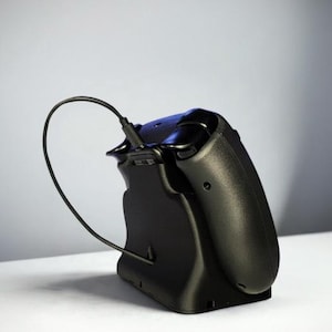 Op de afbeelding: Een zwarte videogamecontroller die in een bijpassend zwart oplaadstation rust. Een dunne zwarte oplaadkabel is in de bovenkant van de controller gestoken, waardoor een gebogen lus ontstaat die verbonden is met de basis van het station.