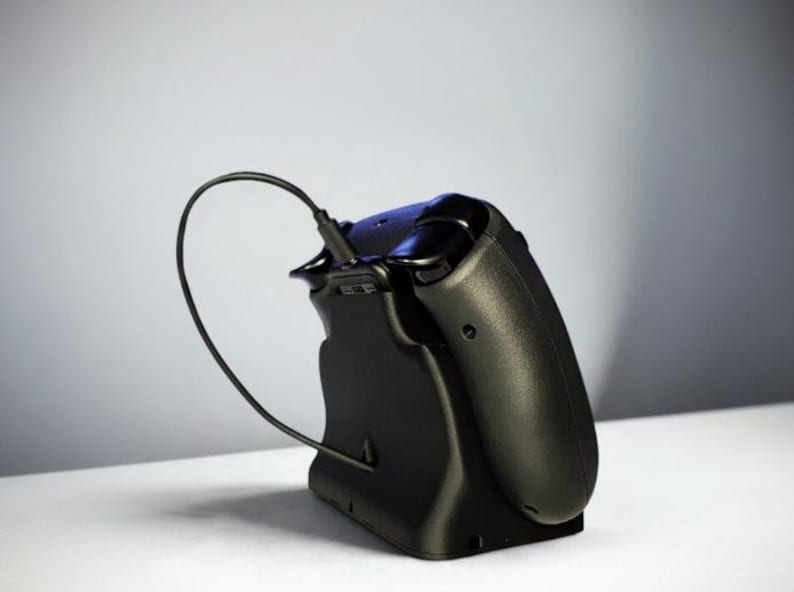 Op de afbeelding: Een zwarte videogamecontroller die in een bijpassend zwart oplaadstation rust. Een dunne zwarte oplaadkabel is in de bovenkant van de controller gestoken, waardoor een gebogen lus ontstaat die verbonden is met de basis van het station.