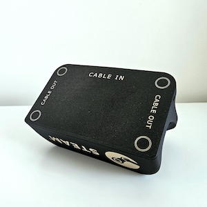 Op de afbeelding: Een zwart rechthoekig elektronisch accessoire met een getextureerd oppervlak met de tekst CABLE IN en CABLE OUT in het wit gedrukt. Het apparaat toont het woord STEAM en een cirkelvormig logo in het wit aan de zijkant.