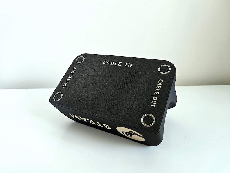 Op de afbeelding: Een zwart rechthoekig elektronisch accessoire met een getextureerd oppervlak met de tekst CABLE IN en CABLE OUT in het wit gedrukt. Het apparaat toont het woord STEAM en een cirkelvormig logo in het wit aan de zijkant.