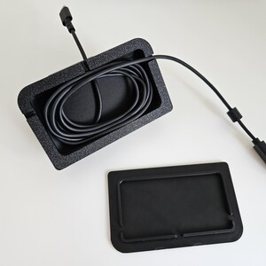 Op de afbeelding: Een zwarte rechthoekige kabelmanagementbak met een getextureerde afwerking, met daarin een opgerolde zwarte USB-kabel met connector. Naast de bak ligt een apart, plat zwart rechthoekig afdekstuk met opstaande randen dat op de bak past.