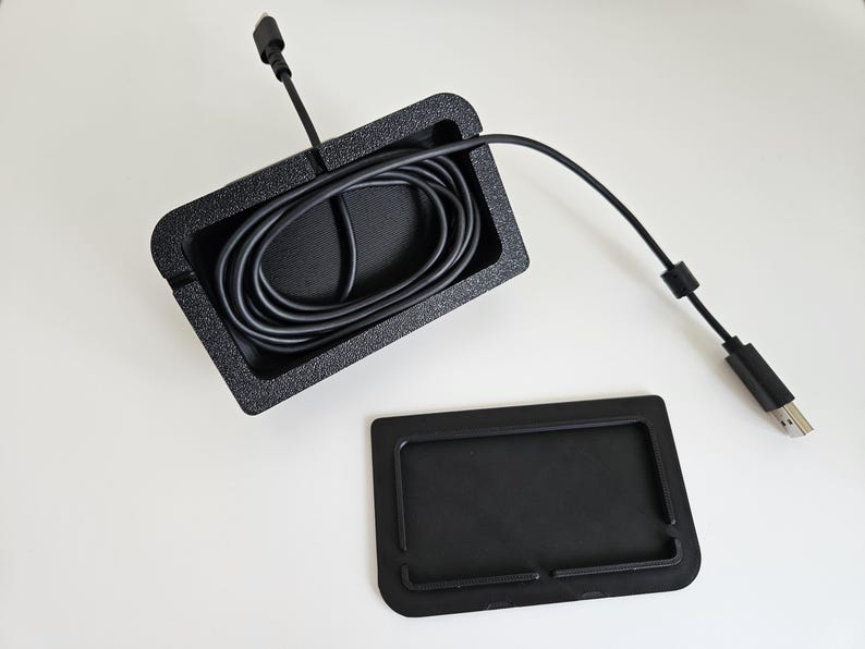 Op de afbeelding: Een zwarte rechthoekige kabelmanagementbak met een getextureerde afwerking, met daarin een opgerolde zwarte USB-kabel met connector. Naast de bak ligt een apart, plat zwart rechthoekig afdekstuk met opstaande randen dat op de bak past.