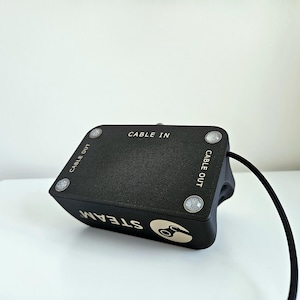 Op de afbeelding: Een zwart rechthoekig elektronisch apparaat met een getextureerd bovenoppervlak met vier zilverkleurige ronde accenten. De tekst CABLE IN en CABLE OUT staat op de bovenkant. De zijkant toont het woord STEAM met een logo en een zwarte kabel.