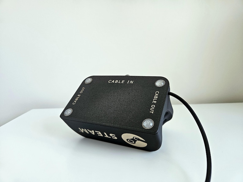 Op de afbeelding: Een zwart rechthoekig elektronisch apparaat met een getextureerd bovenoppervlak met vier zilverkleurige ronde accenten. De tekst CABLE IN en CABLE OUT staat op de bovenkant. De zijkant toont het woord STEAM met een logo en een zwarte kabel.