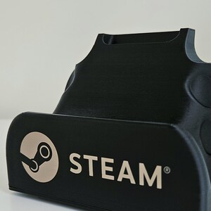 Op de afbeelding: Een zwarte kunststof standaard voor een draagbaar spelapparaat met het Steam-logo en het woord STEAM in een lichtbeige kleur op de voorzijde. De standaard heeft een gebogen, gevormde vorm met een verzonken gleuf aan de bovenkant en ronde inkepingen.