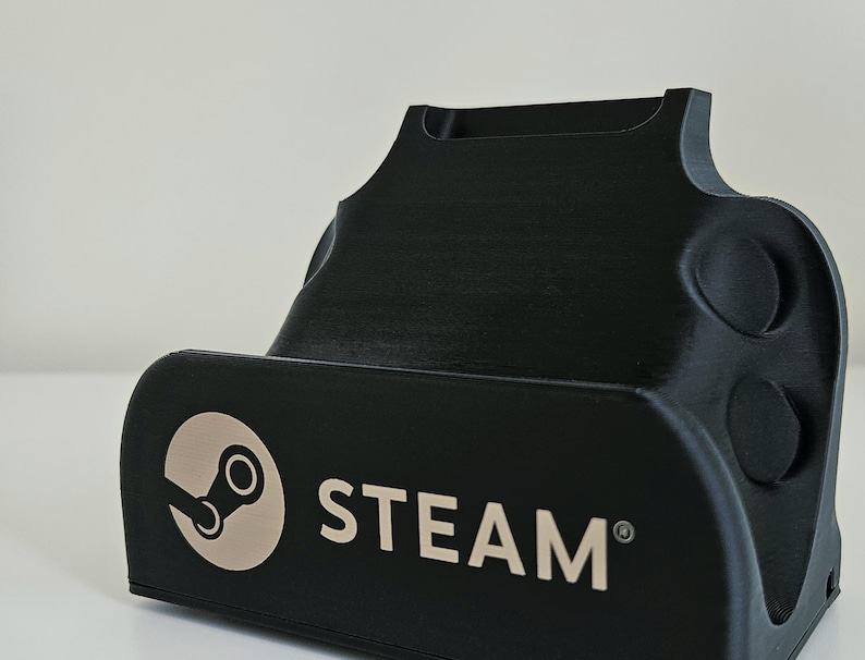 Op de afbeelding: Een zwarte kunststof standaard voor een draagbaar spelapparaat met het Steam-logo en het woord STEAM in een lichtbeige kleur op de voorzijde. De standaard heeft een gebogen, gevormde vorm met een verzonken gleuf aan de bovenkant en ronde inkepingen.