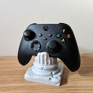 May include: A black Xbox controller rests on a gray, textured stand. The controller has colored buttons and two joysticks. The stand is designed to look like a classical column.